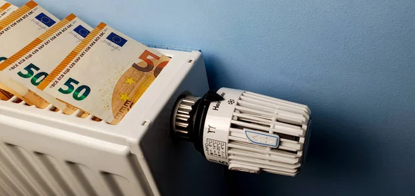 A heating radiator battery with a temperature controller from which 50 euro bills stick out. The concept of high heating costs. Rise in energy prices, energy crisis and recession