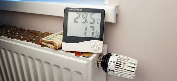 The thermometer that indicates the high temperature is located on the heating radiator battery with a thermostat. The concept of high cost of heating and rational use of resources. A bundle of banknotes of 50 euros and a lot of coins lies on the batt