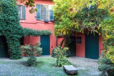 The inner courtyard of the house with a red house wall overgrown with green plants and a bench in the middle of the yard