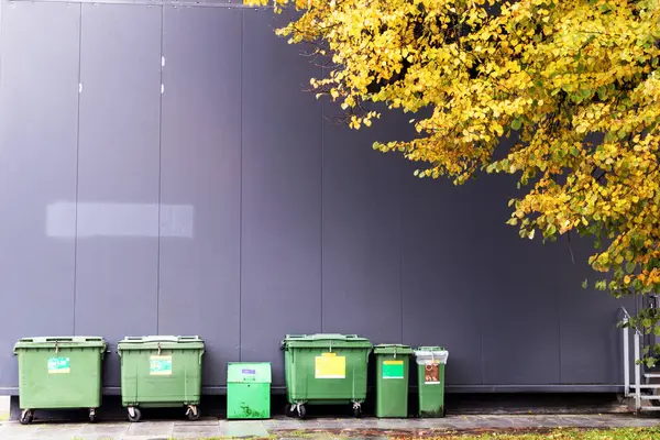 Green waste containers are placed against a gray wall and yellow tree foliage on the side