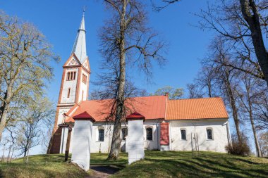 Tepedeki kırmızı kiremit çatılı beyaz kilise açık mavi arka plandaki yeşil çimlerin üzerindeki ağaçların arasında.