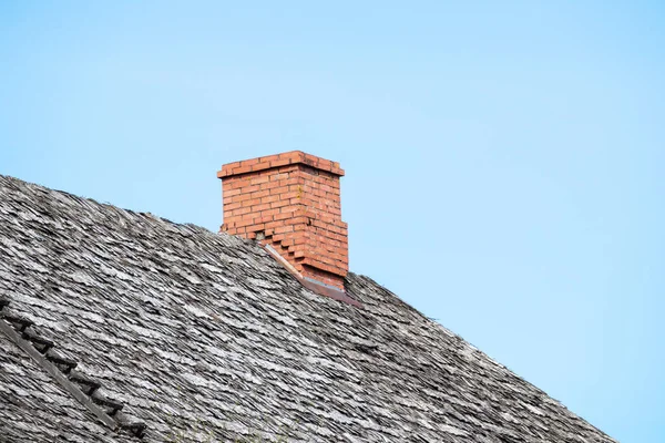 corner of a shingle roof with a ladder and a red brick chimney against a blue sky background