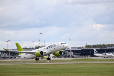 Bir Airbus A220 uçağı havaalanı pistinden kalkışa hazırlanırken, arka planda diğer uçak ve havaalanı binaları yer almaktadır..