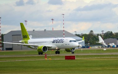 Bir Airbus A220 uçağı havaalanı pistinden kalkışa hazırlanırken, arka planda diğer uçak ve havaalanı binaları yer almaktadır..