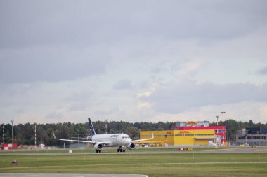 Havaalanı pistinden kalkmaya hazırlanan bir uçak. Arka planda havaalanı binaları ve ağaçlar var..