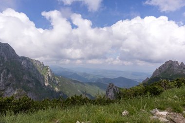 Geniş panoramik bir dağ manzarası. Yeşil yamaçlar, sivri tepeler ve gökyüzünde yüzen beyaz bulutlar..