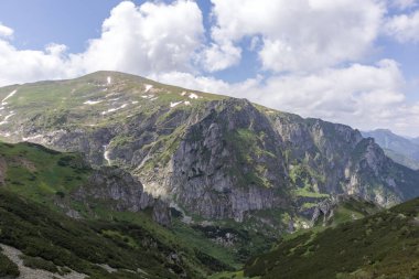 Sivri tepeleri kısmen kar ve yeşil yamaçlarla kaplı dağlık bir bölge, gökyüzünde beyaz bulutlar görülebilir..