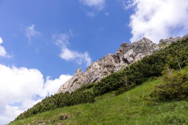 Uçsuz bucaksız yamaçlar. Çimlerle ve çalılarla kaplı kayalık yüzey. Arka planda beyaz bulutlar olan mavi gökyüzü..