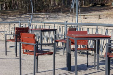 Beton bir zemine yerleştirilmiş birkaç kırmızı masa ve sandalyeli boş bir meydan. Arka planda görünen ağaçlar..