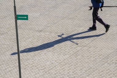 Elinde sopalarla asfalt bir yolda yürüyen yeşil bir levhayı takip eden ve uzun bir gölge bırakan bir adam..