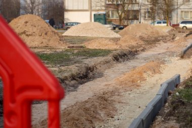 Kazılı bir çukur, birkaç kum yığını ve ön planda kırmızı plastik bariyer olan bir inşaat alanı..
