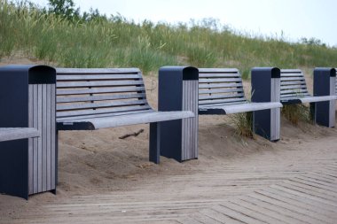 Birkaç ahşap bankta çöp tenekeleri sıralanmış kumlu bir plajın arka planına karşı..