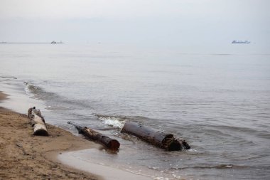 a seashore with several metal pipes washed out of the water, ships in the background. clipart