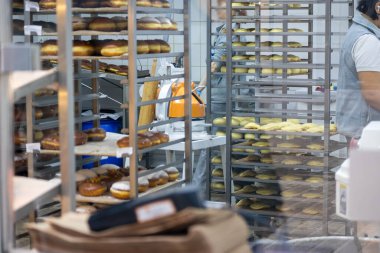 Metal raflarda çok sayıda donut görülebiliyor ve cam bir vitrinden görülebiliyor. Garson sırtı seyirciye dönük duruyor..