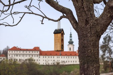 a birdcage hanging from a tree and a large building with a red roof and a white tower can be seen in the distance. clipart