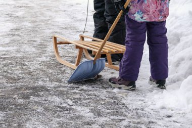 İki kişi arka planda bir kürek ve tahta bir kızakla kar temizliyor..
