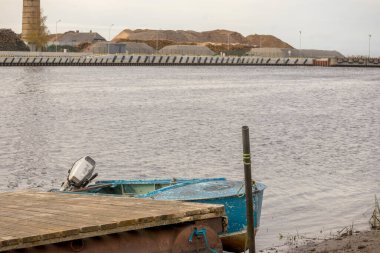 Mavi tekne rıhtımda, arka planda sanayi yığınları var..