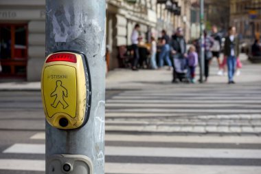 Yaya geçidinde beyaz ve gri çizgili bir yaya geçidinde bulunan direğin üzerinde bir yaya trafik ışığı düğmesi, kaldırımda insanlar ve arka planda görünen bir bina.