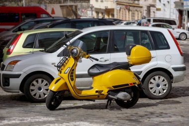Şehirdeki bir otoparkta birkaç arabanın arasına park etmiş sarı bir scooter..