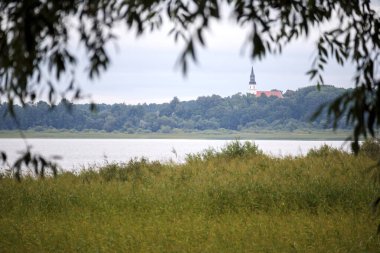 Önünde çimenlik bir kıyısı, arkasında uzanan sakin bir su kütlesi ve sivri kulesi olan yeşil bir orman ile bulanık ağaç dalları arasında manzara