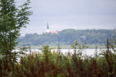 Önünde yeşil ağaçların yayıldığı bir su kütlesi olan bir manzara ve arka planda sivri uçlu bir kuleye, muhtemelen bir kiliseye sahip bir bina görülebilir..