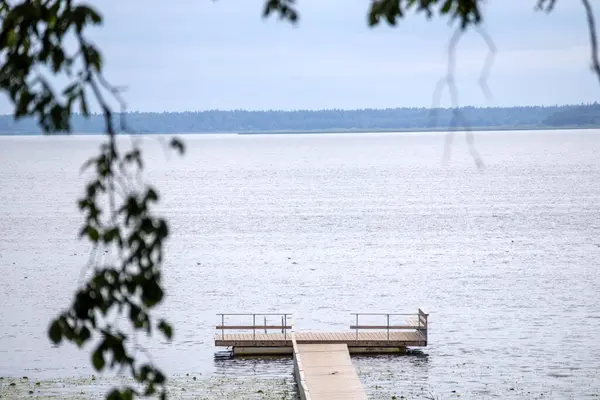 Sakin suya uzanan ahşap bir iskele, sonunda küçük bir platform ve bulutlu bir gökyüzünün altında bulanık ağaç dalları..