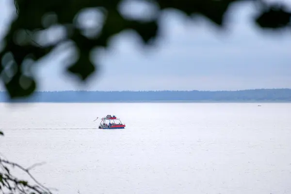 Uzakta sakin bir su kütlesi üzerinde yüzen küçük, kırmızı ve beyaz bir tekne, ön planda bulanık siyah ağaç dalları ile.