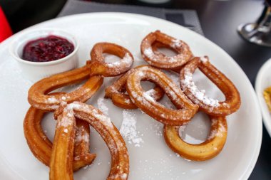 Bir tabak altın sarısı, üzerine pudra şekeri serpiştirilmiş churros ve koyu kırmızı soslu küçük beyaz bir tabak, belki reçel veya böğürtlen sosu.