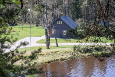 Mavi çatılı, küçük, karanlık bir evin manzarası. Suya yakın ağaçlık bir alanda. Ön planda ağaç dalları görülebilir, bu da görüntünün bitki örtüsü yoluyla çekildiğini gösterir..