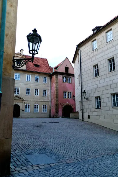 Şehirde sessiz, kaldırımlı bir meydan. Meydan çeşitli renkler ve mimari tarzlara sahip binalarla çevrilidir. Sol tarafta kırmızı çatılı ve duvarında siyah fener olan parlak bir bina var.