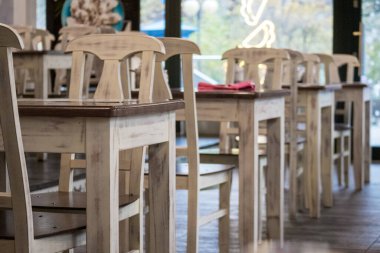 Sıraya dizilmiş birkaç açık ahşap masa ve sandalyeli bir kafe ya da restoran alanı ve arka planda pencereleri olan ışıklı bir duvar, rahat ve ferah bir atmosfer yaratıyor..