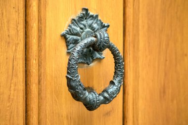 Açık kahverengi ahşap kapıya bağlı, halka şeklinde bir kapı kolu olan dekoratif, koyu metal bir kapı tokmağı görülür..