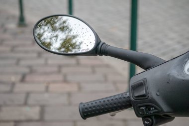 Yeşil ağaçları ve gökyüzünü yansıtan damlalar halinde yüzeyi olan bir motosiklet ya da scooter dikiz aynası. Arka planda asfalt bir yol var..