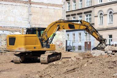 Büyük, sarı bir kazıcı, arka planda eski binalar ve kum ve moloz yığınları bulunan bir inşaat sahasında bulunuyor..