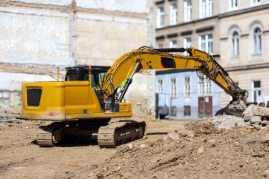 Bir inşaat sahasında büyük, sarı bir kazıcı, eski bina temellerinin yakınında ya da yıkılmış bir bina ve şehir binalarının zemininin karşısında yer alıyor..