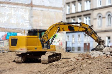 Büyük, sarı bir kazıcı molozlarla dolu bir inşaat sahasında duruyor ve arka planda eski şehir binaları görünüyor..