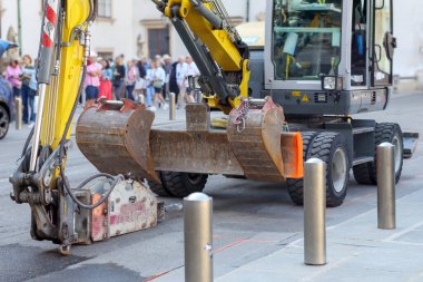 A yellowish excavator with a bucket and hydraulic equipment standing on the street near a sidewalk with metal poles, with blurred people and city buildings visible in the background.