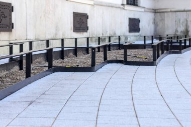 Modern, gri kaldırım taşı yürüyüş sistemi. Siyah metal kenarlı ve parmaklıklı. Dekoratif pencereli beyaz bina duvarı boyunca uzanan..