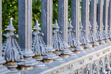 Dekoratif, koyu metal çitler, sivri yüzeyli ve süslemeli elementler, yeşil ağaçlar ve bulanık arka planda çalılar..