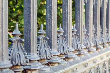 Dekoratif, koyu metal çitler, sivri yüzeyli ve süslemeli elementler, yeşil ağaçlar ve bulanık arka planda çalılar..