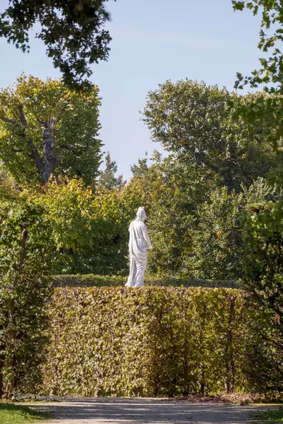 açık mavi gökyüzünün altındaki ağaçlarla çevrili yeşil bir çitin üzerinde duran beyaz bir heykel..