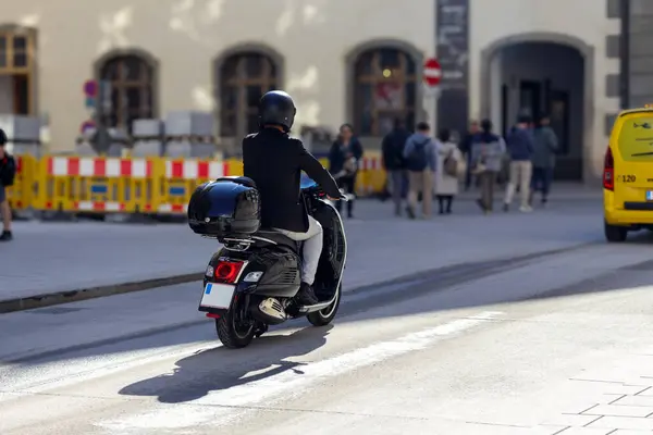 Arkasında bagaj kutusu olan siyah bir scooter kullanan biri, arka planda insanlar ve binalarla şehir caddesinde ilerliyor..