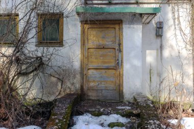 Sarı ahşap kapısı ve iki penceresi olan eski, yıpranmış bir binanın girişi. Kısmen sarmaşıklarla kaplı ve merdivenlerin altında kar kalıntıları var..