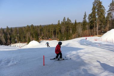 Karlı bir kayak yamacı, yamaçtan aşağı inen iki kayakçı, arka planda bir orman ve kar yığınları var..