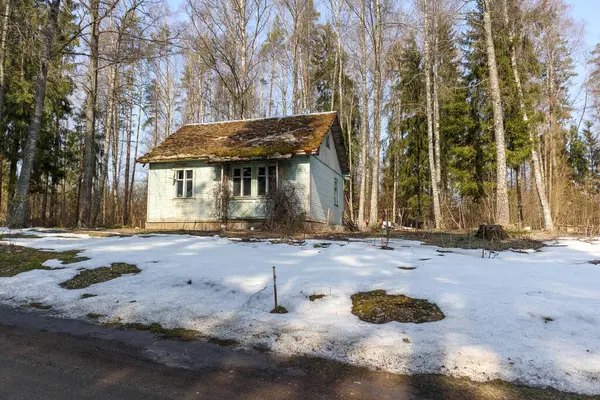 Ormanın kenarında kahverengi çatılı, küçük, eski, açık mavi bir ev ve önünde eriyen kar parçalarıyla kaplı bir arazi var..