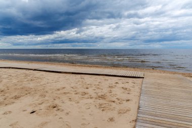 Bulutlu bir gökyüzünün altında, koyu mavi bir denize uzanan ahşap patikaları olan kumlu bir sahil..