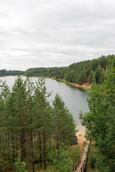 Kumlu, sakin bir göl manzarası, bulutlu bir gökyüzünün altında geniş yeşil bir çam ormanıyla çevrili. Tahta bir patika suya doğru iniyor..