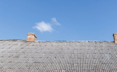 Tuğla bacalı, yıpranmış gri çatılı açık mavi gökyüzüne karşı kırsal mimariyi tasvir eden ince bir bulutla belirgin bir şekilde duruyor..