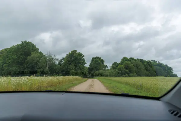 Bir arabanın bakış açısından, toprak bir yol yeşil tarlalar ve ağaçlar arasında kıvrılır, eski bir taş duvar görünür, bulutlu bir gökyüzünün altında.