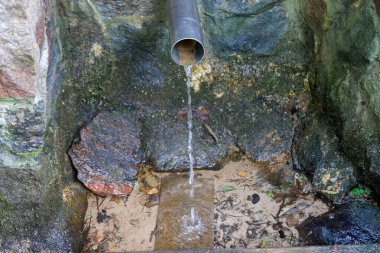 Temiz su, metal bir borudan yosunlu kayalar ve kumla çevrili doğal bir bahar havzasına akar, saflığı ve doğayı sembolize eder..